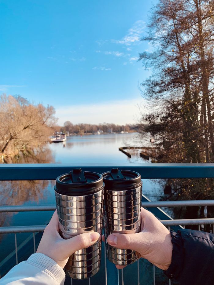 Services-01 Two people holding reusable mugs by a serene lake on a sunny day.