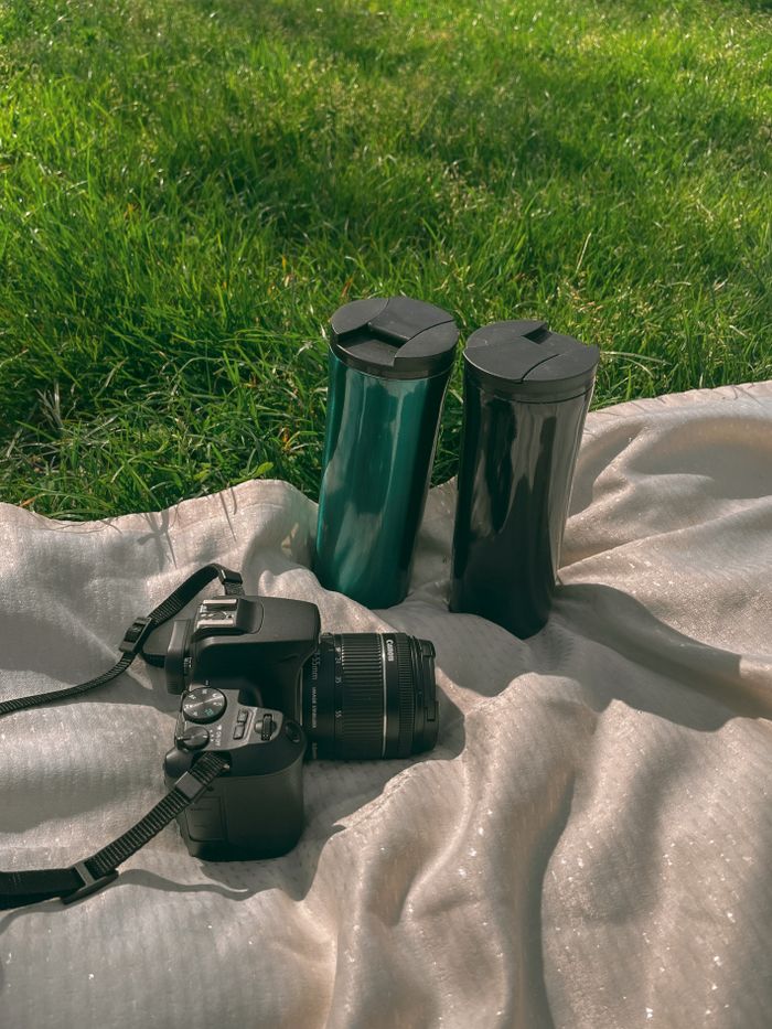 creative A DSLR camera and two coffee mugs on a blanket outdoors in green grass.