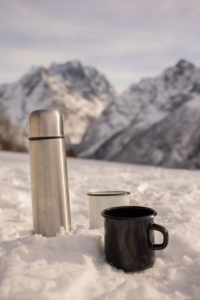 Thermos and mugs in snow with snowy mountain backdrop for a winter adventure.