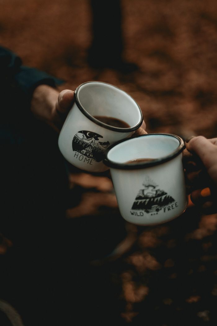 Services-02 Two people toasting with enamel mugs around a campfire in a forest, creating a warm and adventurous vibe.